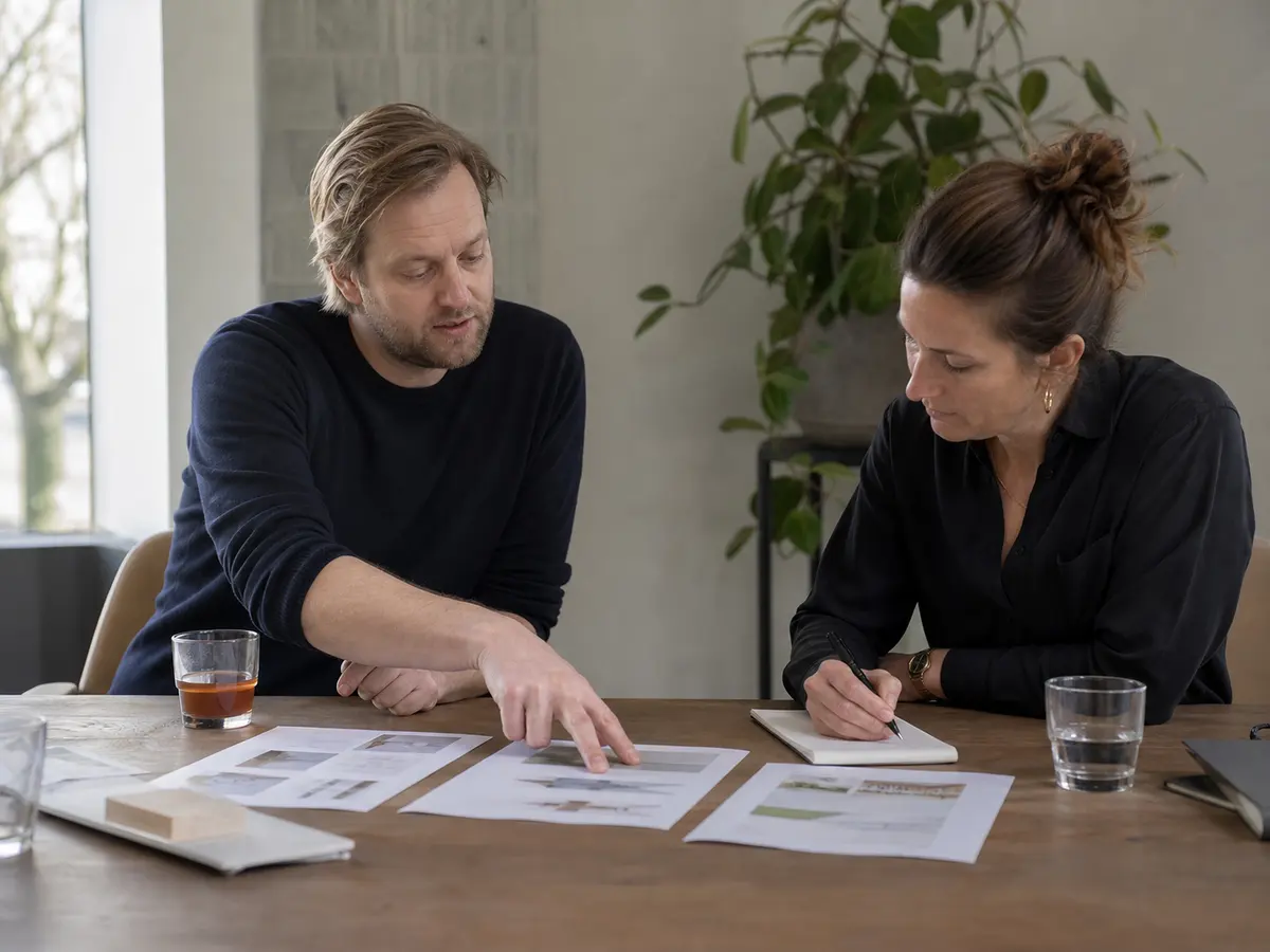 Twee mensen zitten aan een tafel met drie geprinte pagina's naast elkaar. De een wijst er een aan, de ander schrijft mee op een blocnote.