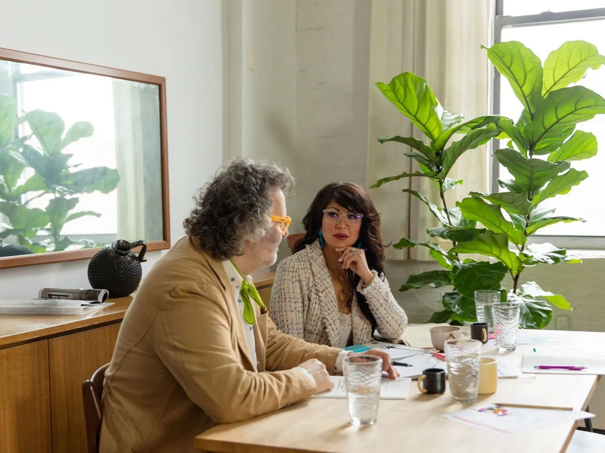 Twee mensen aan een tafel met een laptop en aantekeningen, in gesprek.