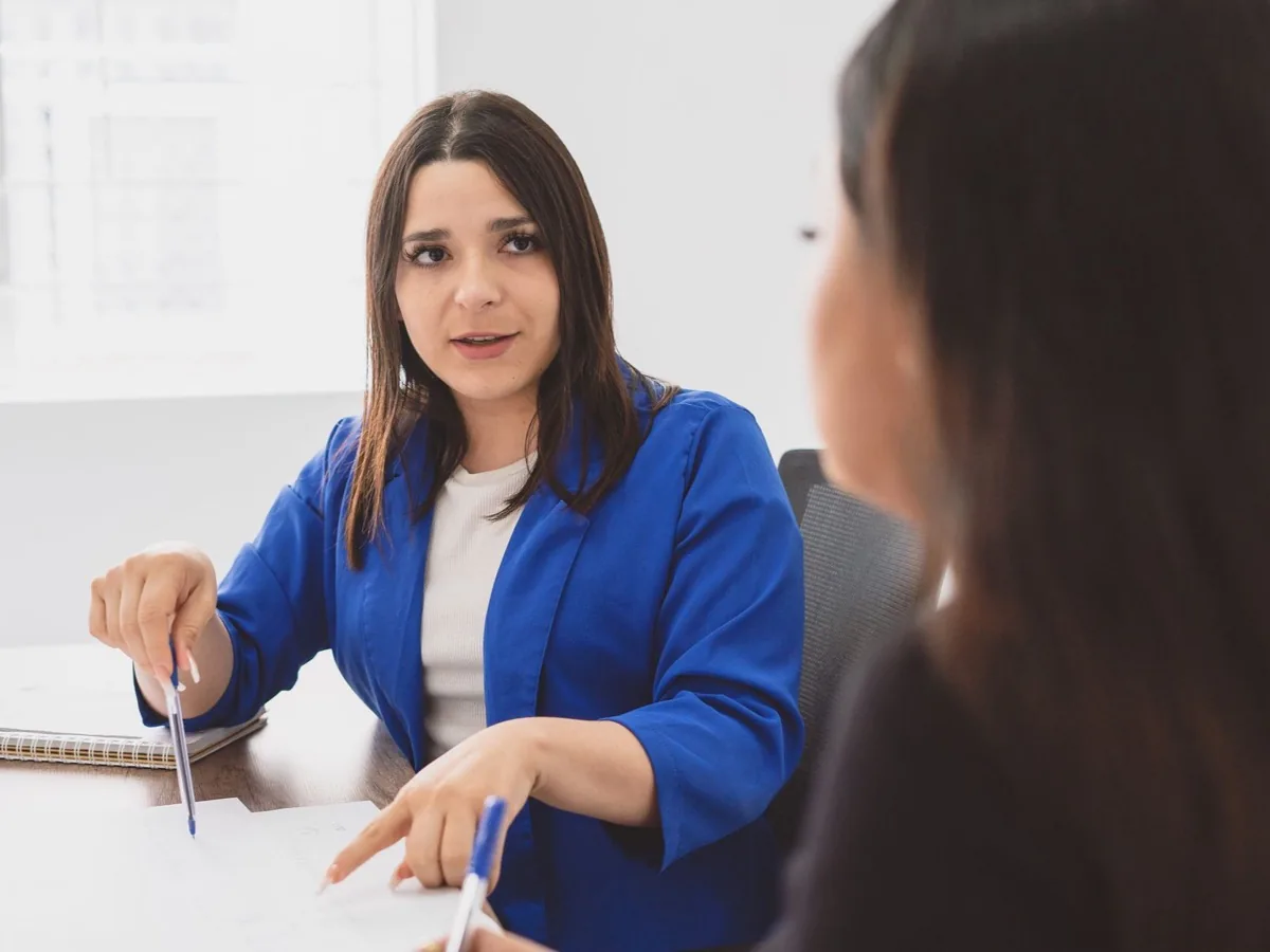 Twee vrouwen in gesprek aan een tafel, een van hen gebaart tijdens het praten.